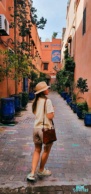 The writer of the post in casual wear and a hat stands in a colorful alleyway in Marrakech, observing the traditional architecture and vibrant street art
