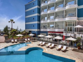 Outdoor pool at Hôtel Club Val d'Anfa in Casablanca, with sun loungers, white parasols and palm trees beside the blue and white hotel facade.