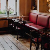 Cozy restaurant interior with red upholstered bench seating, dark wooden tables and chairs, wooden floor, and a window with potted plants.