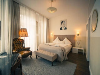 A neatly made bed with pillows, a tufted bench, an armchair, a desk, two nightstands, lamps, and sheer curtains in a softly lit hotel room.