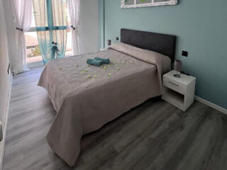 A neatly made bed with a gray headboard and taupe bedding, beside a white nightstand, in a modern bedroom with wood floors and floor-to-ceiling windows.