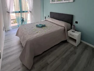 A neatly made bed with a gray headboard and taupe bedding, beside a white nightstand, in a modern bedroom with wood floors and floor-to-ceiling windows.