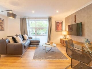 Living room at Merchant City Glasgow Green Apartment, with a brown leather corner sofa, arc floor lamp, round coffee table and a wall-mounted TV beside a glass desk.