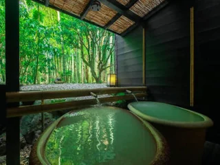 Two soaking tubs filled with water sit in a sheltered area with a bamboo roof, overlooking a lush green bamboo forest through an open wall.
