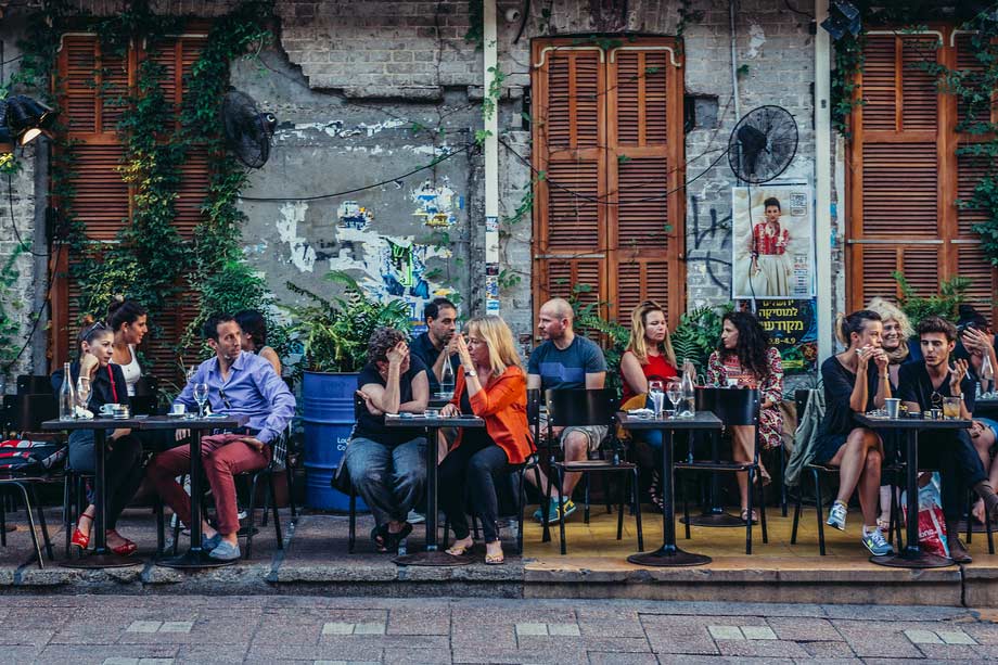 A lively outdoor café scene with patrons enjoying drinks and conversation at tables set along a street with rustic charm and greenery