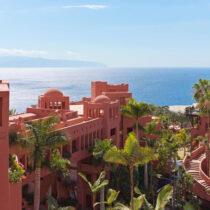 Coral-colored resort buildings with domed rooftops, surrounded by palm trees, overlook the ocean under a clear blue sky.