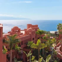 Coral-colored resort buildings with domed rooftops, surrounded by palm trees, overlook the ocean under a clear blue sky.