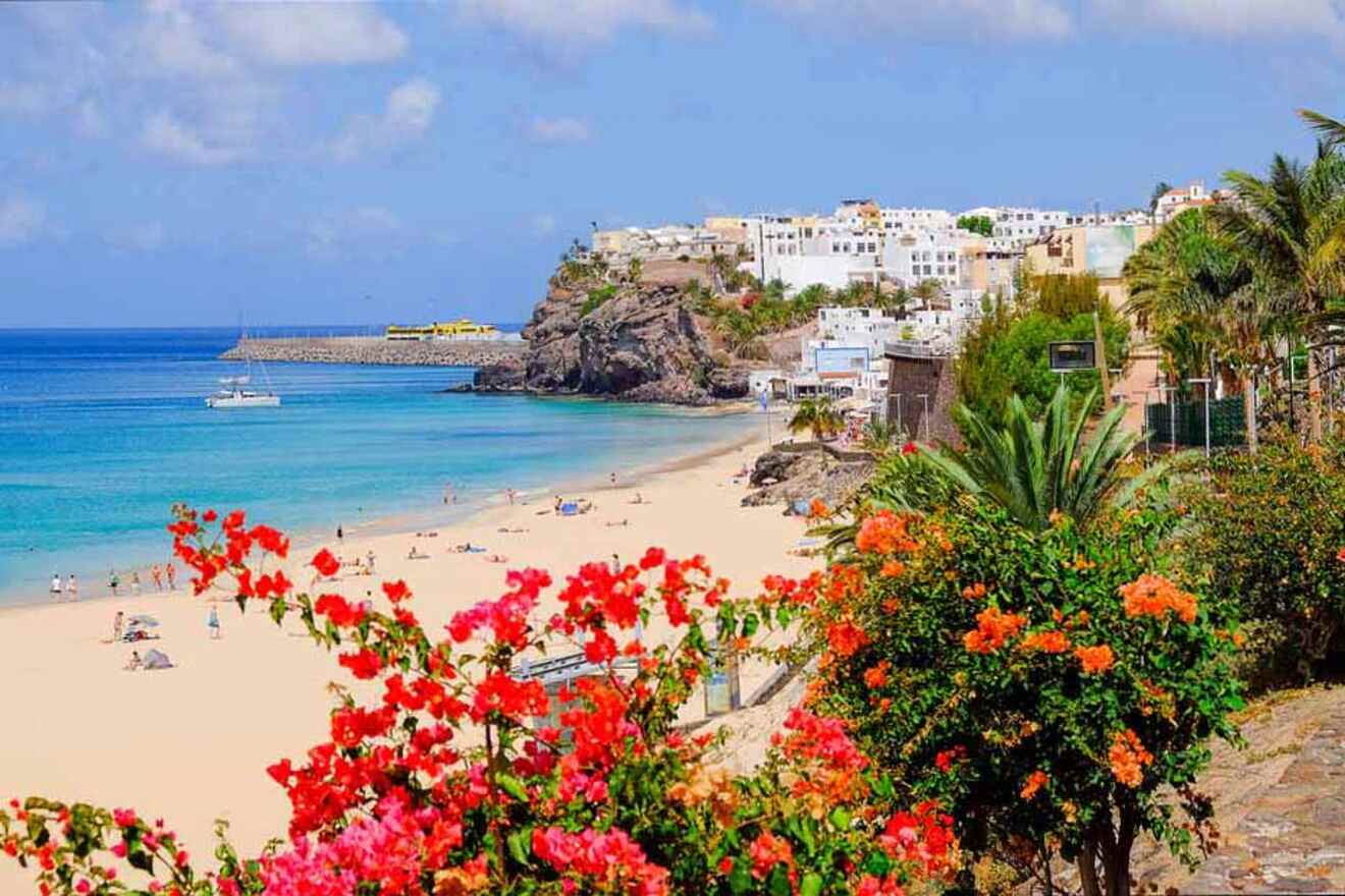 Sandy beach with crystal-clear waters in Fuerteventura, lined with colorful flowers and a cliffside white village