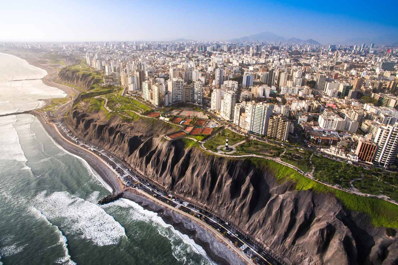 Aerial view of a coastal city next to steep cliffs with a beach and breaking waves in the foreground.