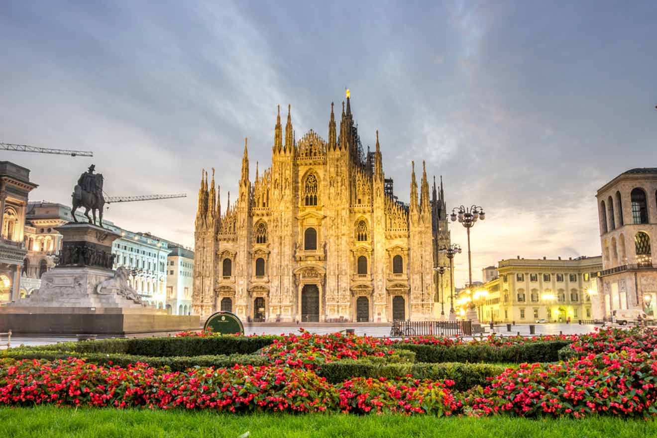 Milan cathedral (duomo di milano) with surrounding architecture and blooming red flowers at dusk.