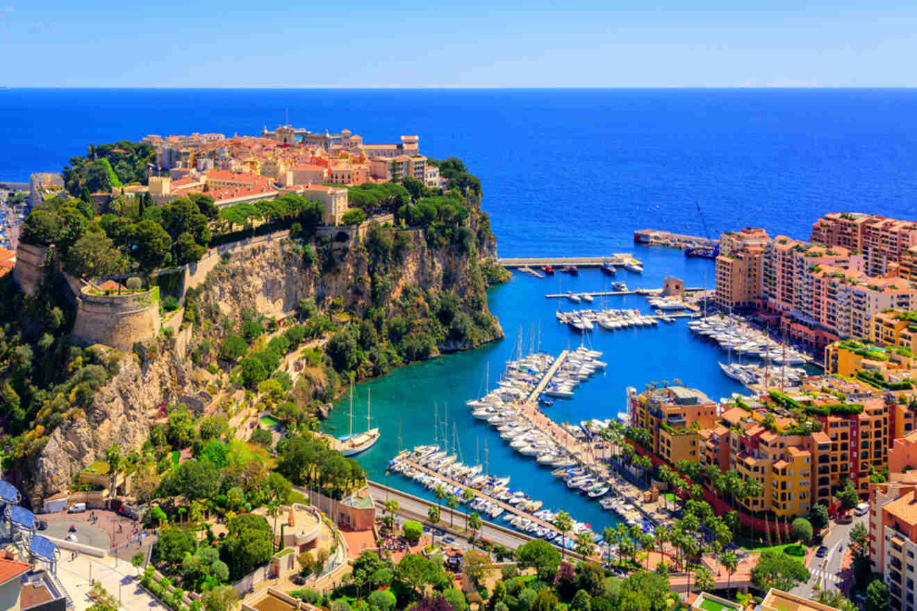 Aerial view of Monaco, showcasing the vibrant architecture with terracotta roofs, the historic Prince's Palace, lush greenery, and the Monaco Harbour filled with yachts on a bright sunny day