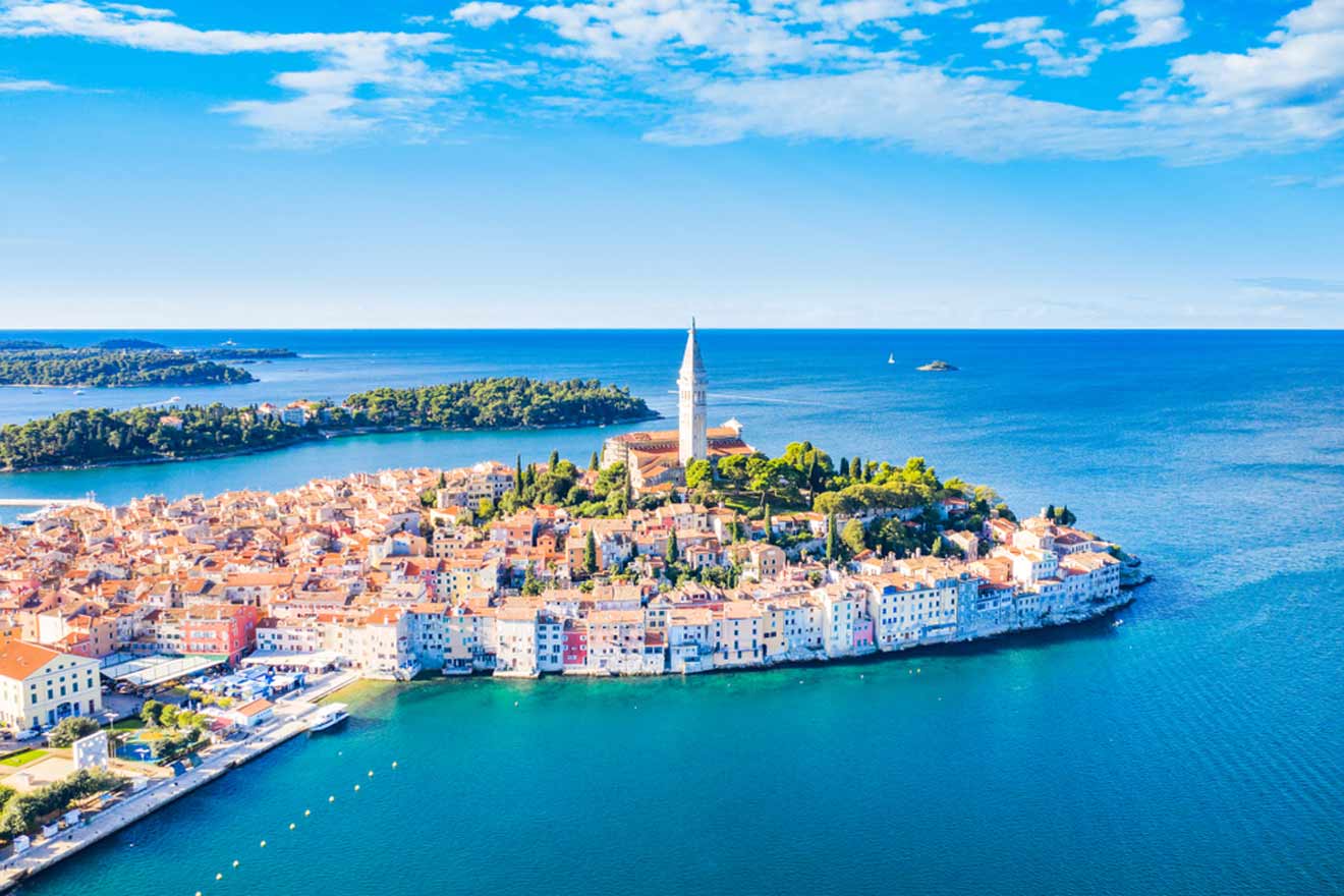 Aerial view of Rovinj, Croatia, showcasing the coastal old town with terracotta rooftops and a prominent bell tower, surrounded by the bright blue Adriatic Sea with boats and a green peninsula in the background