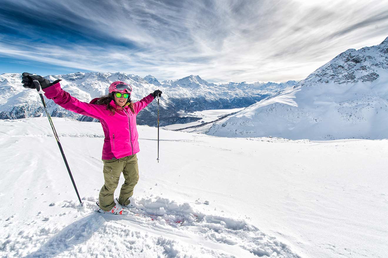 Joyful skier in a bright pink jacket and goggles, arms raised in triumph, stands on a snow-covered slope with a stunning panoramic view of the Swiss Alps behind her