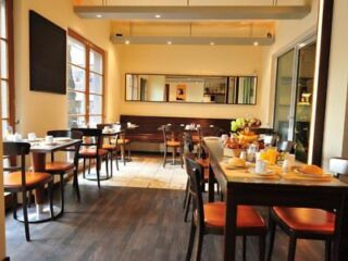 Small restaurant dining area with neatly set tables, wooden chairs, large windows, and a buffet with breakfast items in the background.