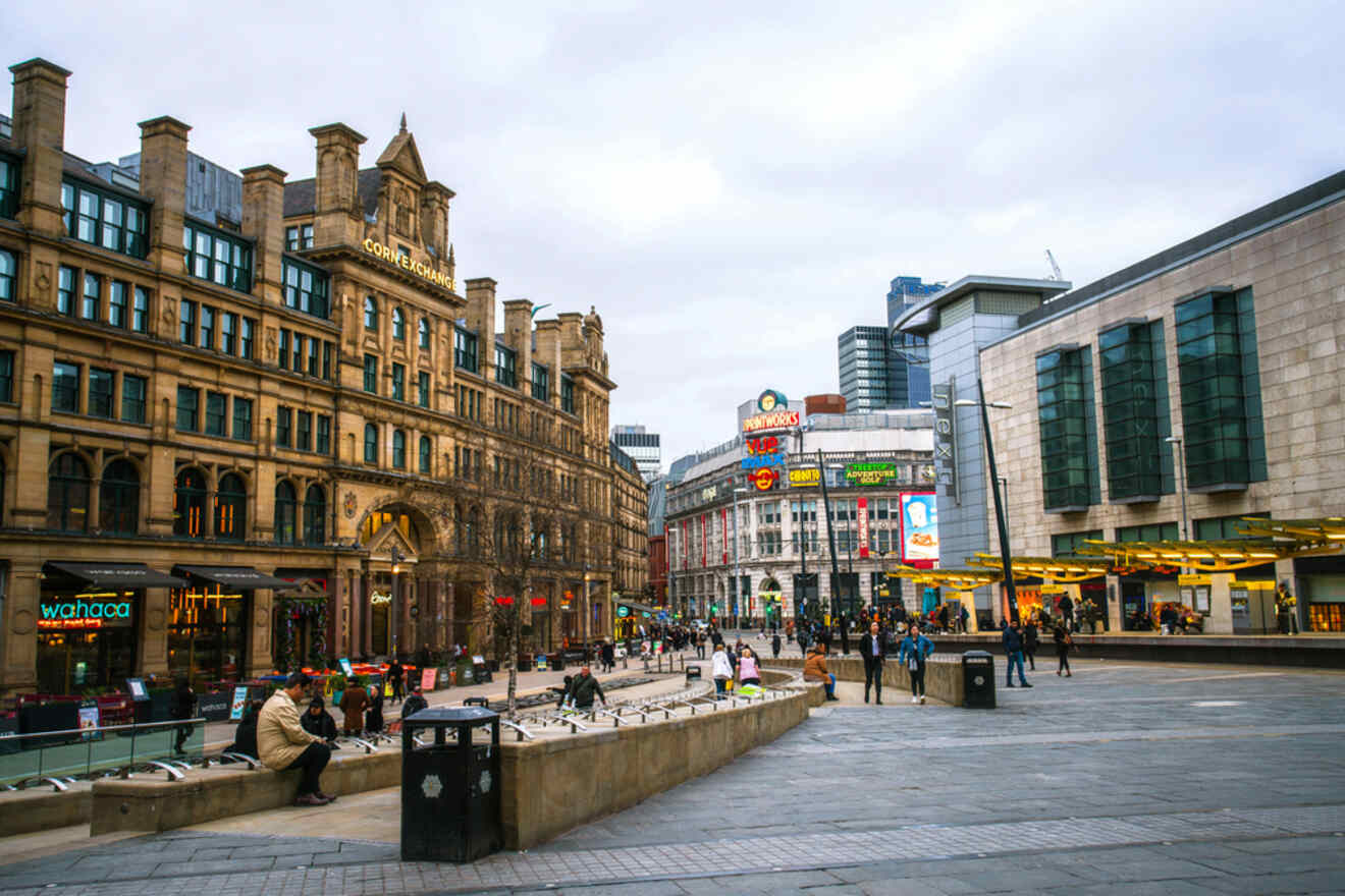 A bustling square in Manchester with the historic Corn Exchange building on the left and modern storefronts, against a backdrop of city architecture and grey skies