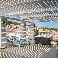 Rooftop terrace with striped overhead pergola casting shadows on white furnishings and loungers, overlooking a coastal town.