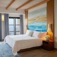 Un dormitorio moderno con una cama grande, ropa de cama blanca, dos mesitas de noche con lámparas, un cuadro con temática del océano azul y una ventana con cortinas azules con vistas al mar.