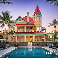 Victorian-style mansion with a turret and wrap-around porch next to a pool at dusk