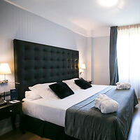 A neatly made king-size bed with white linens and black pillows in a modern bedroom with gray curtains and side lamps.