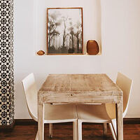 Rustic dining area with a worn wooden table, white chairs, and a framed painting on a patterned wall.