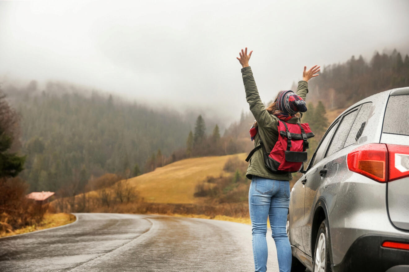 A female traveler with a red backpack raises her hands joyfully next to a parked car on a misty road amidst a scenic mountainous landscape