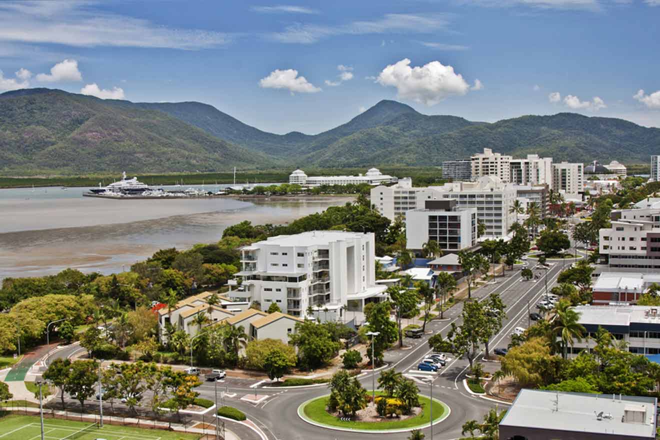 Aerial view of a Cairns with modern buildings, a wide river, lush greenery, and mountains in the background on a clear day.