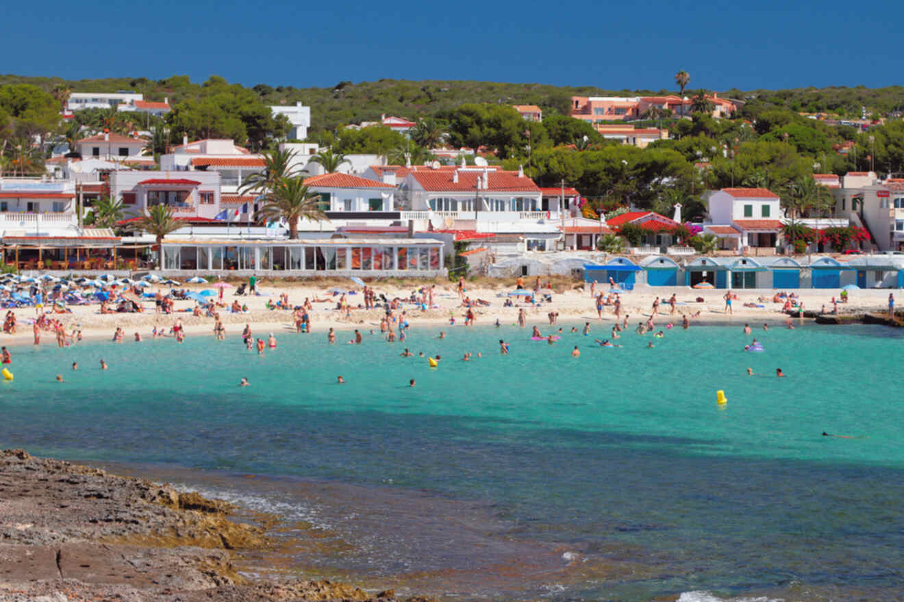 A bustling beach scene in Punta Prima with people swimming in clear turquoise waters, sandy shore lined with colorful beach umbrellas, and a vibrant collection of white-washed buildings in the background