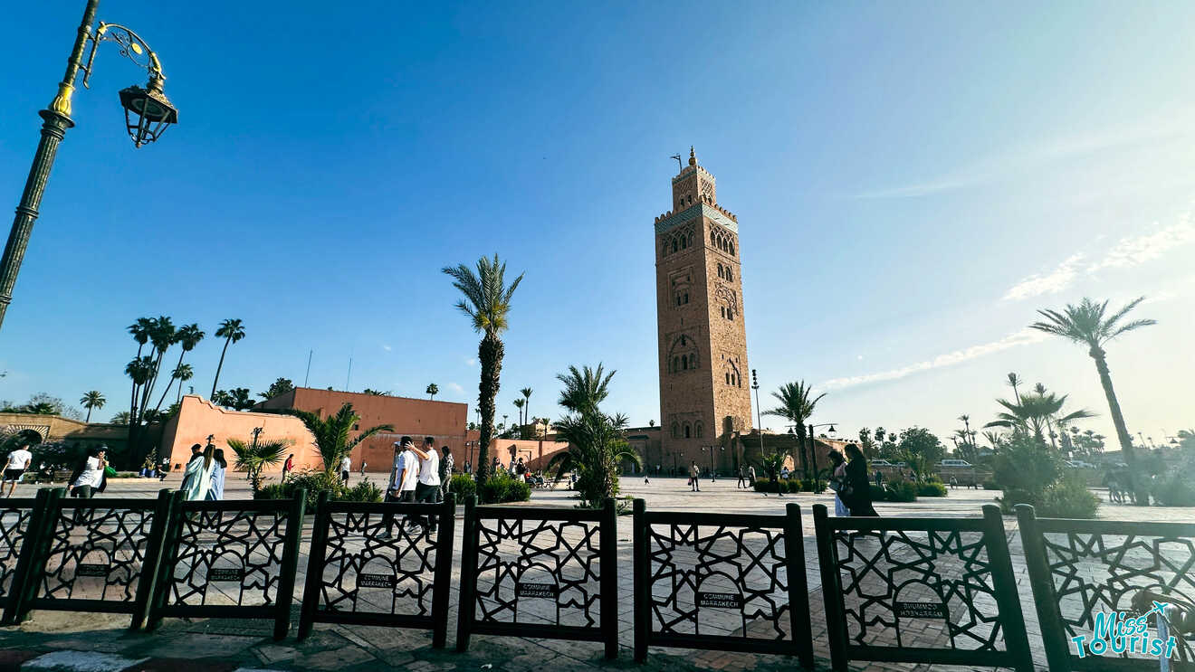 A bustling square in the Northern Medina of Marrakech with the iconic Koutoubia Mosque in the background, pedestrians, and palm trees under a clear blue sky