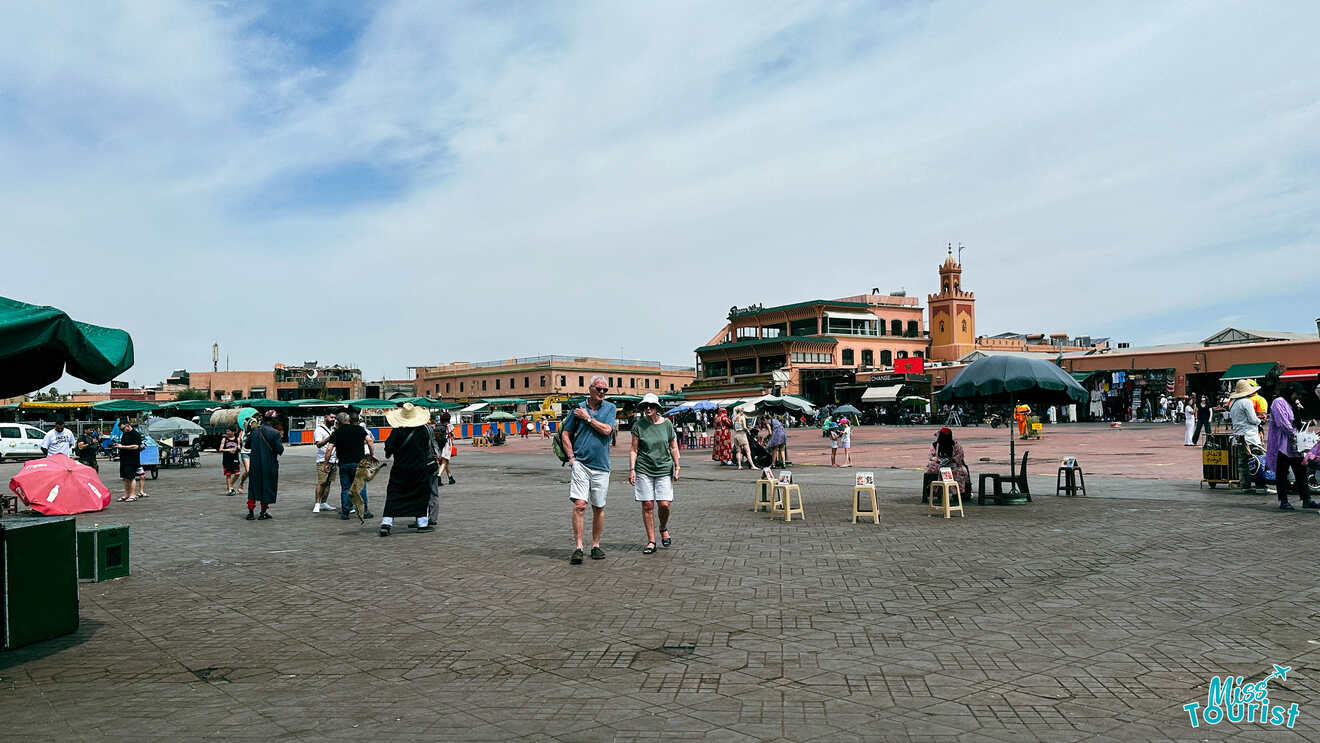 Visitors walking across the lively Jemaa el-Fnaa market square, surrounded by market stalls and the distinct architecture of Marrakech under a bright sky