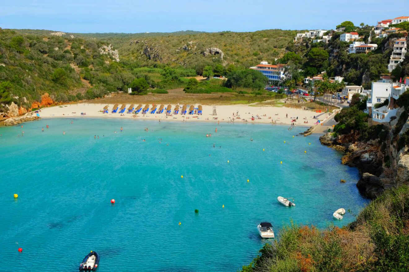 The inviting cove of Cala en Porter, Menorca, with a sandy beach, loungers waiting for sunbathers, and crystal-clear water, bordered by rugged cliffs and greenery