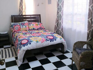 A small, bright bedroom with a colorful floral-patterned bedspread, checkered black and white floor, sheer curtains, and a wicker chair.