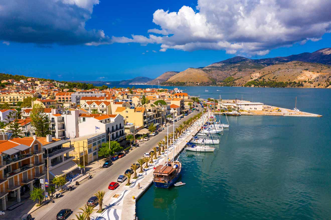 Aerial view of a mediterranean coastal town with a marina lined with boats, colorful buildings, and surrounded by mountains under a clear sky.