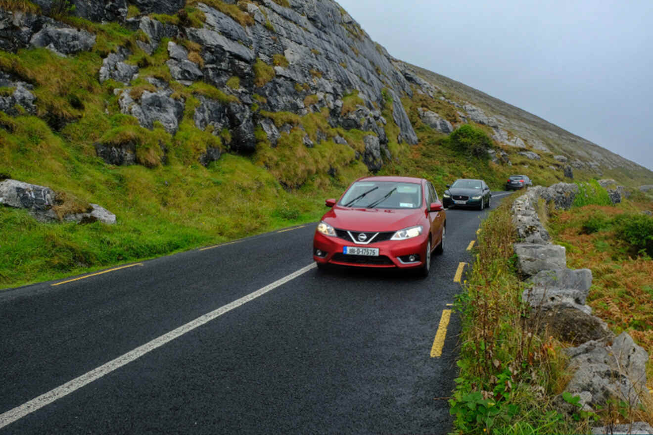 Cars traveling on a winding road through a lush, rocky landscape under overcast skies, capturing the experience of driving in the Irish countryside