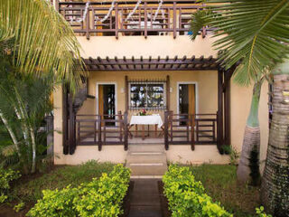 Exterior of a tropical beach house with double doors, a balcony above, surrounded by lush palm trees and shrubs.