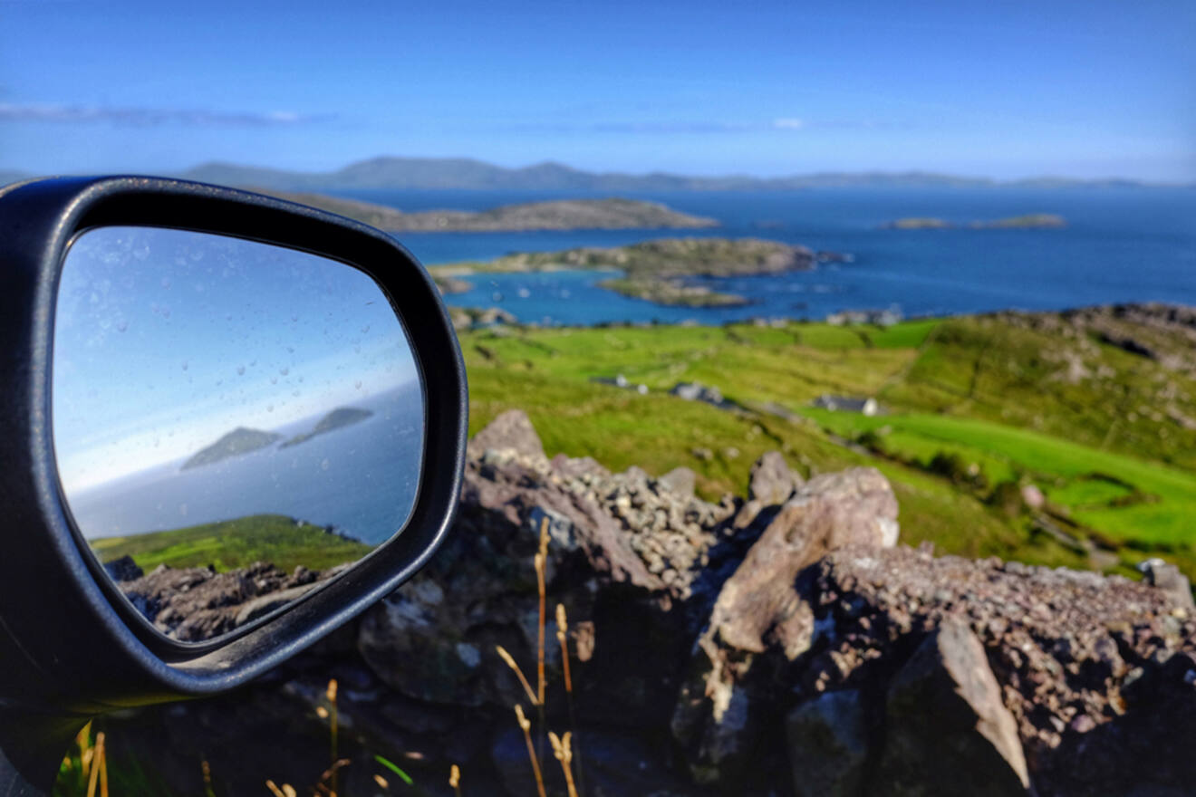 View through a car's side mirror reflecting a stunning landscape with clear blue skies, coastal waters, and green terrain, illustrating the concept of exploring Ireland's scenic routes