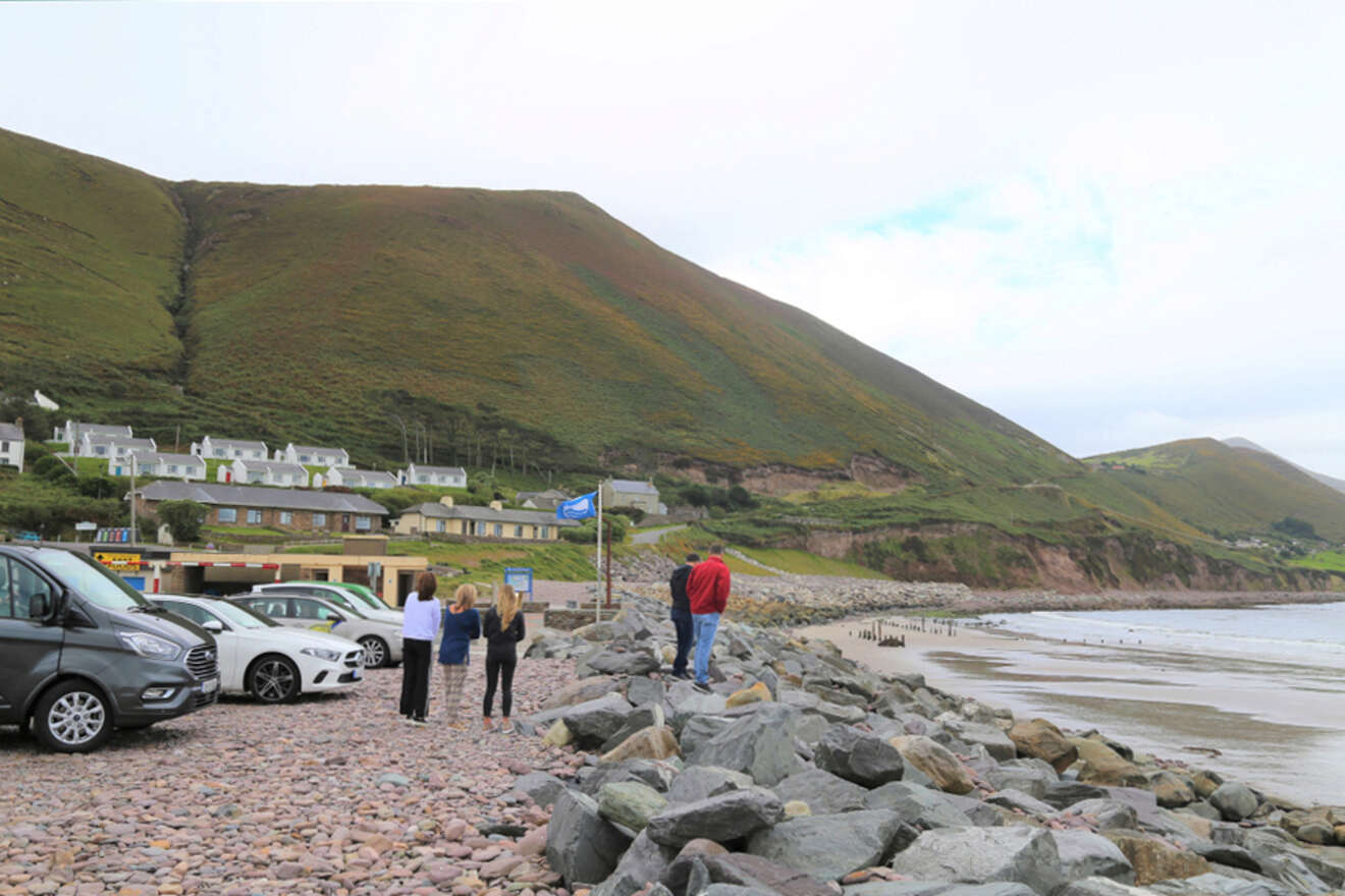Tourists and parked cars on a pebbled beachfront with a view of a green hillside and a quaint village, exemplifying a serene coastal town in Ireland