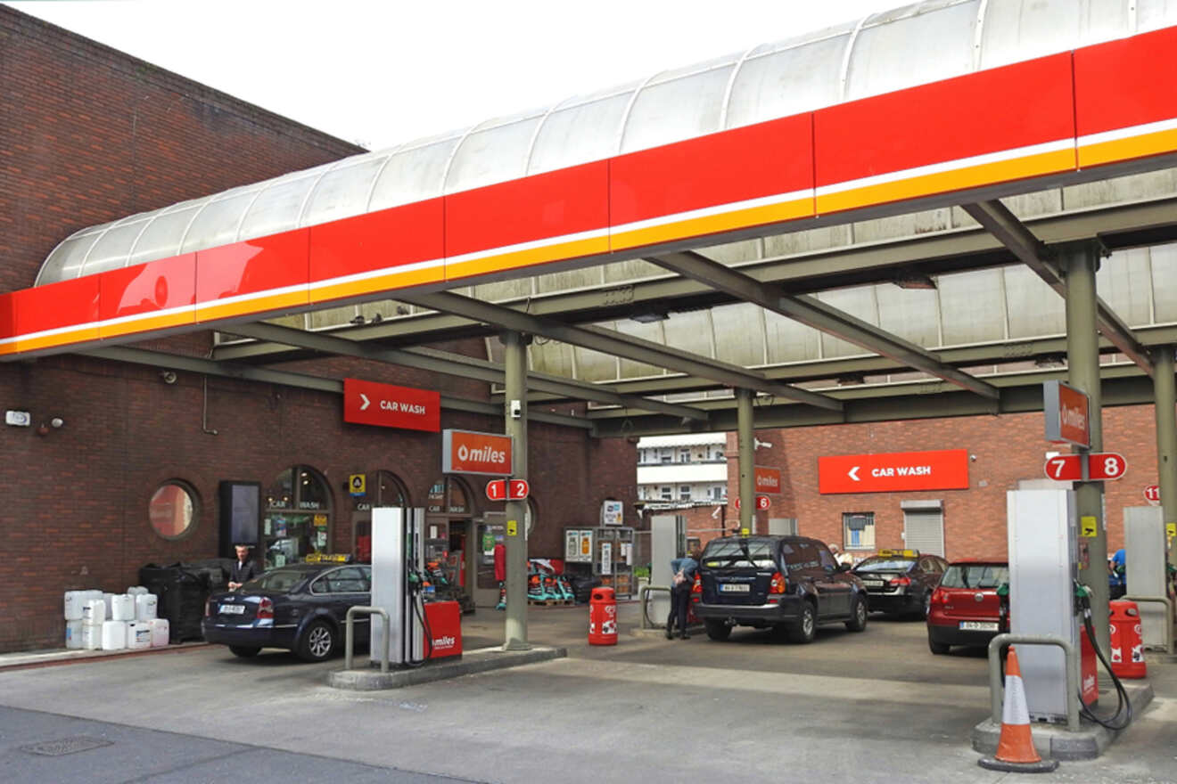 A bustling petrol station with multiple cars refueling under a red and white canopy, adjacent to a car wash, showcasing a typical urban fuel stop