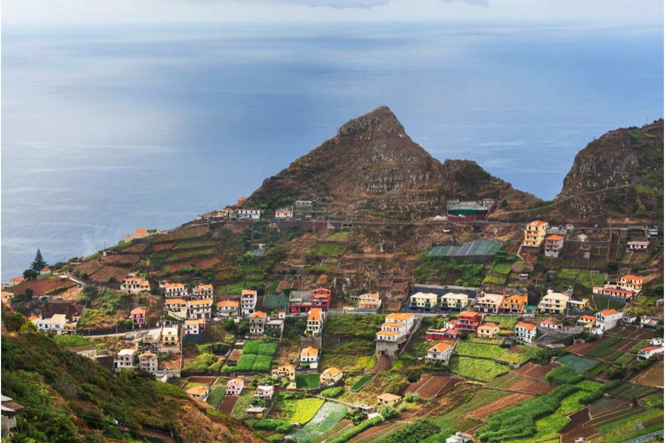 A sweeping view over terraced fields and clusters of houses in Madeira, with a rugged mountain backdrop meeting the vast ocean