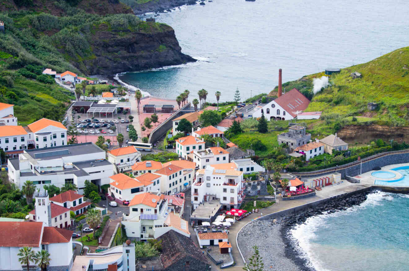 An idyllic coastal town in Madeira, with historical white buildings and red roofs, a church spire, and a natural pool, embraced by the Atlantic Ocean