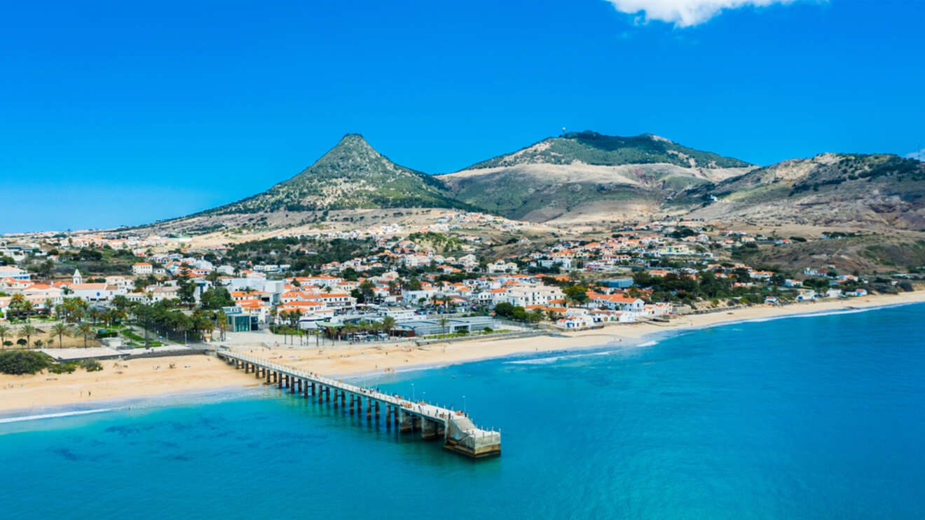 A panoramic view of a sandy beach and pier in Madeira, with a small town against the backdrop of a striking mountain and clear blue skies