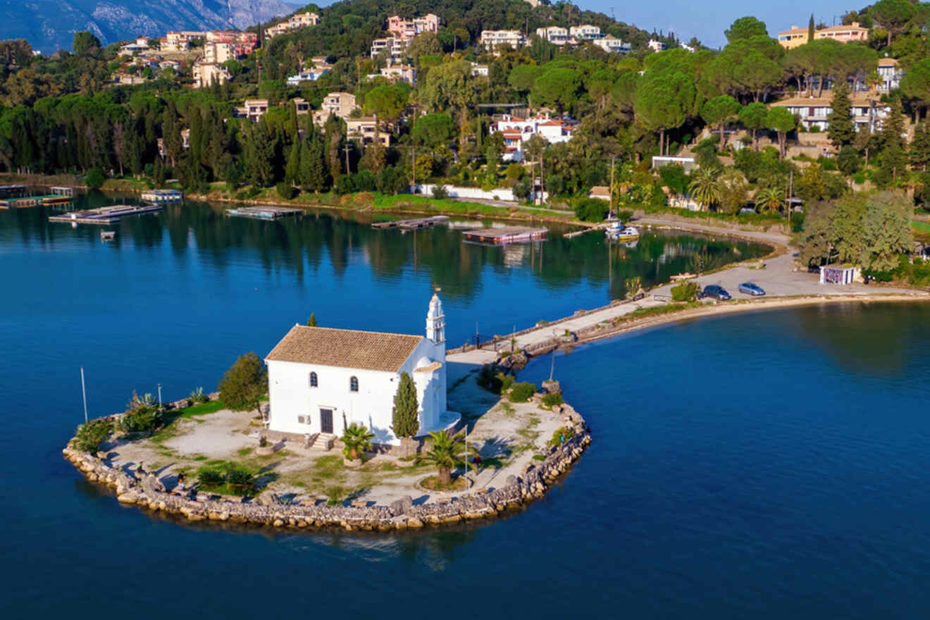 Isolated white chapel with a bell tower on a small islet connected by a narrow causeway, surrounded by calm azure waters and a lush coastal landscape.