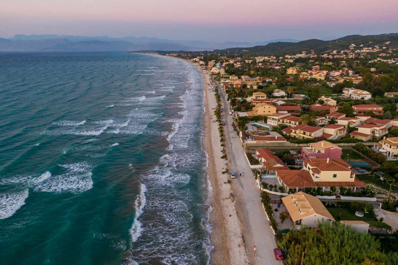Twilight hues settle over a sweeping beach with gentle waves, lined by a coastal road and a series of homes, creating a serene transition from day to night along the seaside.