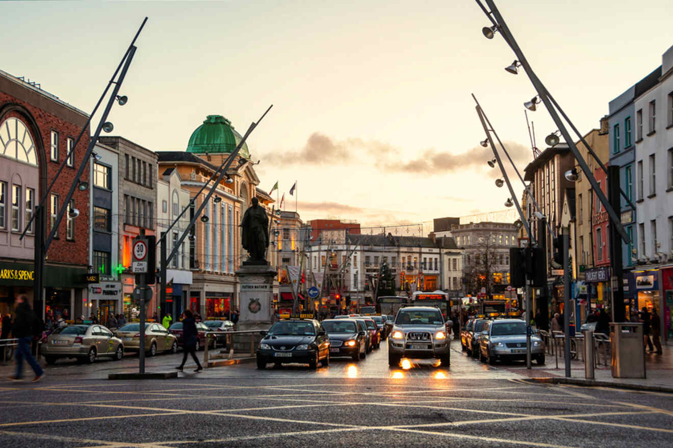Dusk falls on a lively street in an Irish city, where cars queue at a traffic light under the watchful gaze of a statue, with storefronts casting a warm glow onto the wet pavement, encapsulating a vibrant urban evening