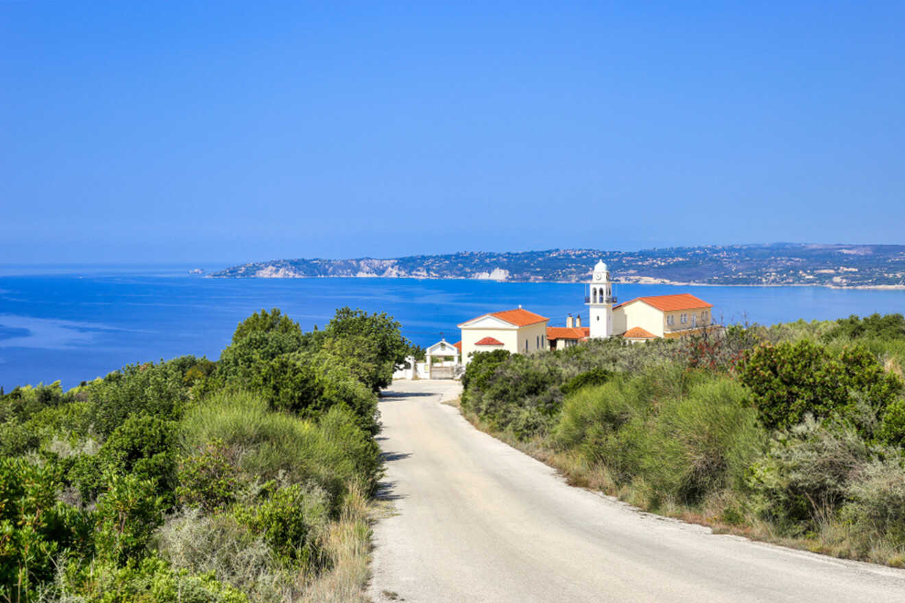 A scenic coastal road leading to a small village with red-roofed buildings and a church, overlooking a clear blue sea and distant cliffs.