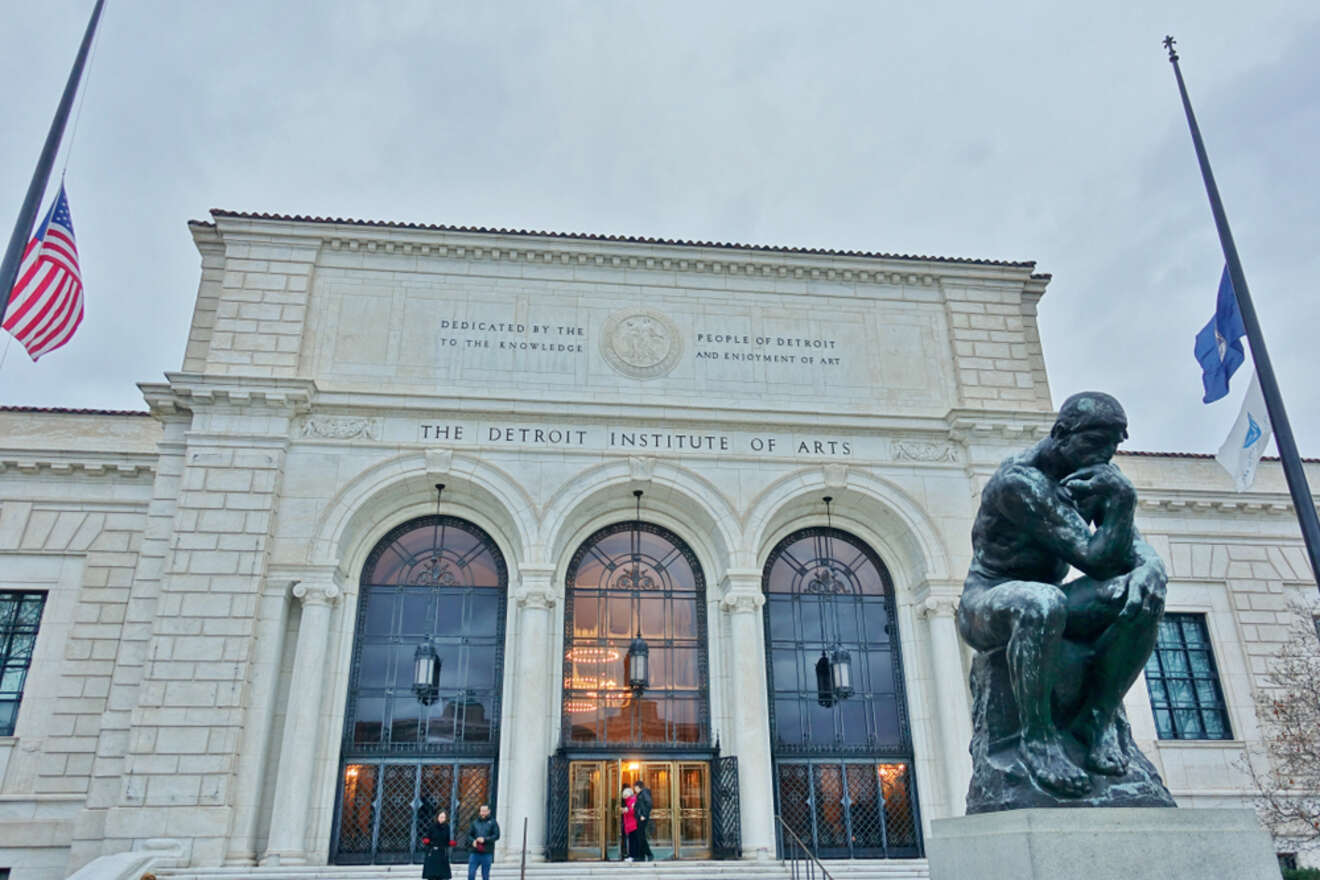 The neoclassical facade of the Detroit Institute of Arts under an overcast sky, with an iconic bronze statue of 'The Thinker' by Rodin in the foreground