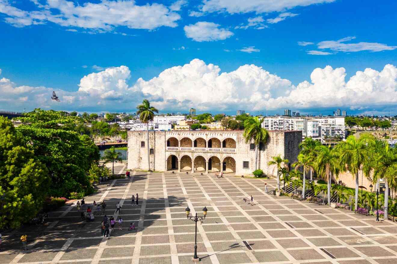 A historical plaza in Santo Domingo with an old fortress, people walking, and a city skyline in the background.