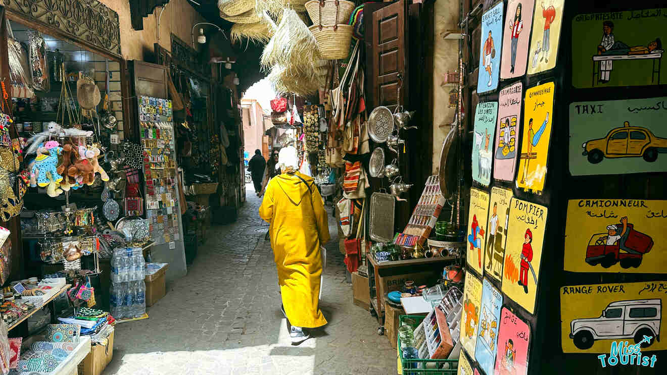 A person in a yellow djellaba walking through a narrow alley of the Southern Medina, flanked by shops selling traditional Moroccan goods