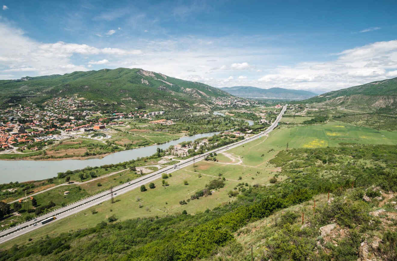 Aerial view of a winding river flowing through a lush valley with a highway parallel to it, flanked by green hills and scattered settlements under a cloudy sky