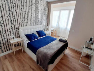A modern bedroom featuring a white double bed with navy blue bedding, birch tree patterned wallpaper, and light hardwood flooring.