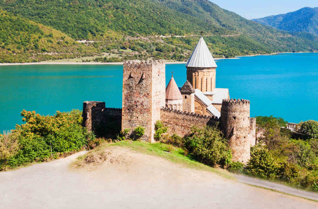 The Ananuri Fortress stands majestically by a turquoise lake, with green forested hills in the background, showcasing historical architecture in a serene natural setting